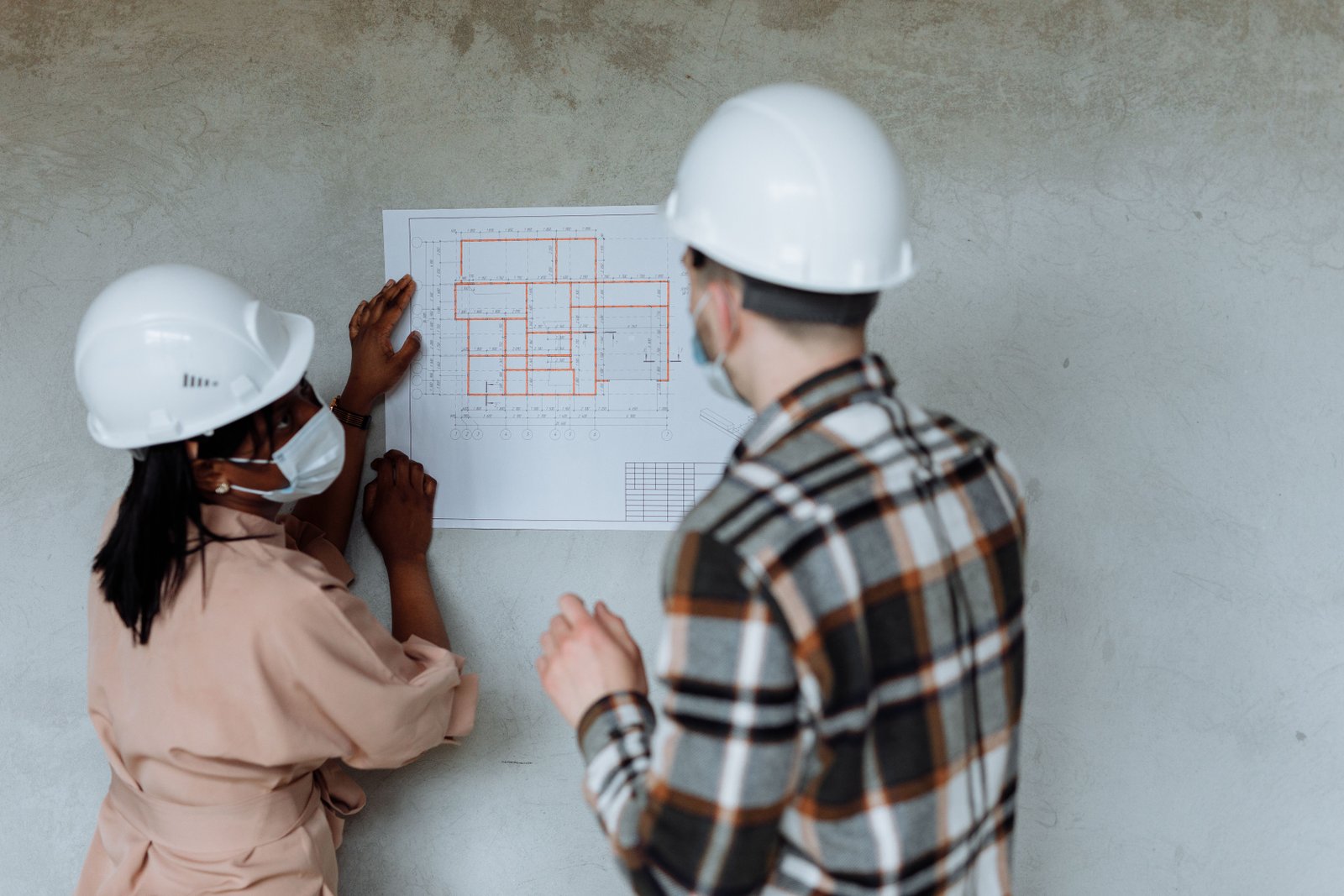 Two professionals in hard hats reviewing an architectural plan displayed on a wall, photographed by Thirdman